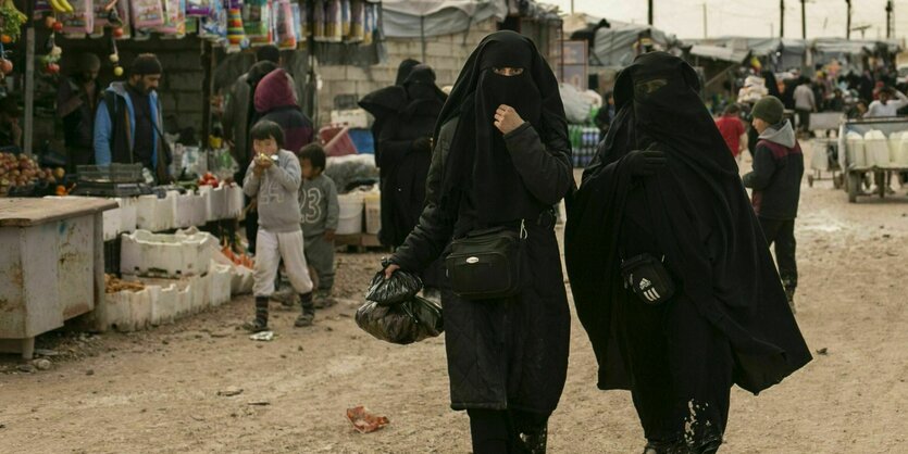 Zwei vollverschleierte Frauen laufen durch ein Camp, links ein Lebensmittelstand