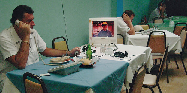 Ein Mann sitzt an einem Restauranttisch und telefoniert. Auf seinem Tisch steht ein alter Computer- oder Fernsehbildschirm.