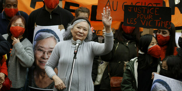 Ältere Frau vor Demonstrierenden spricht in ein Mikrofon und hebt die linke Hand