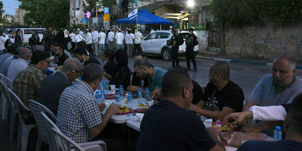 Männer sind in der Dämmerung auf der Straße an einem langen Tisch und essen, im Hintergrund Siedler und Soldaten