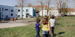 Drei spieldene Kinder auf einer Wiese, hinten sind helle und flache gebäude zu sehen