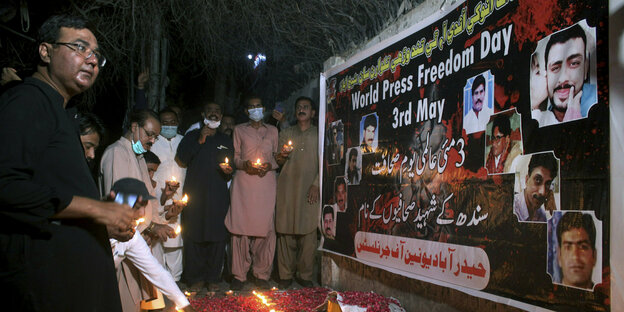 Ein paar Männer stehen vor einem Plakat, davor brennen ein paar Kerzen