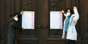 Zwei Frauen stehen vor einem Kircheneingang und hängen Plakate auf