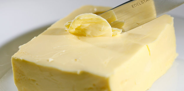 Wir sehen ein Stück Butter auf einem weißen Teller und ein Messer, das sich in die Butter gräbt.