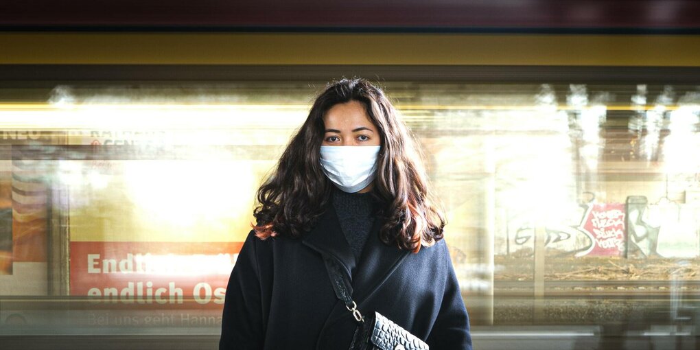 Eine Frau mit Corona-Maske steht in einer U-Bahnstation und blickt in die Kamera