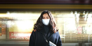 Eine Frau mit Corona-Maske steht in einer U-Bahnstation und blickt in die Kamera