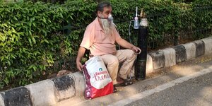 Ein Mann sitzt mit einer Sauerstoffflasche und einer Atemschutzmaske erschöpft am STraßenrand