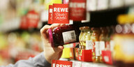 Eine Hand greift ein Glas Marmelade aus einem Supermarktregal, drumherum Schilder auf denen steht "REWE Beste Wahl"