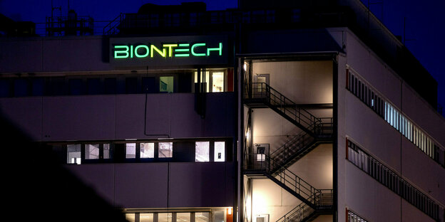 notbeleuchtetes mehrstöckiges Gebäude mit großen Fenstern und grünen Biontech-Leuchtschriftzug auf dem Dach