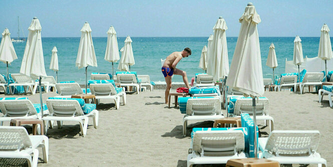 Ein Mann trägt zwischen Strandliegen und Sonnenschrimen mit Blick auf das Meer Sonnencreme auf