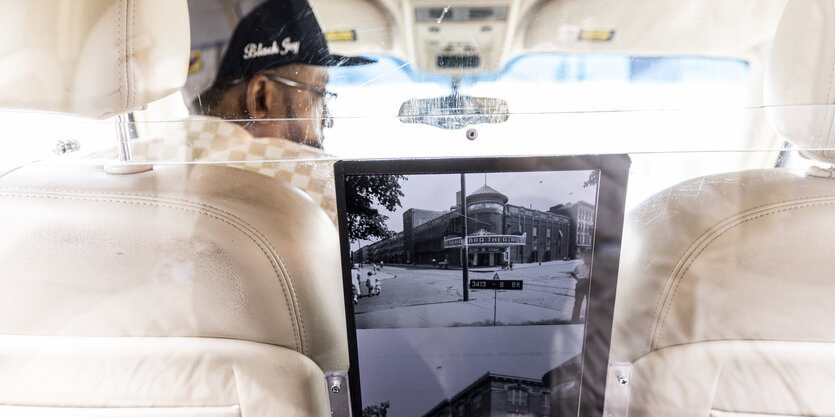 Ein Mann sitzt auf dem Fahrersitz eines Taxis, zwischen ihm und den Rücksitzen sind eine Plexiglasscheibe und zwei Bildschirme platziert.