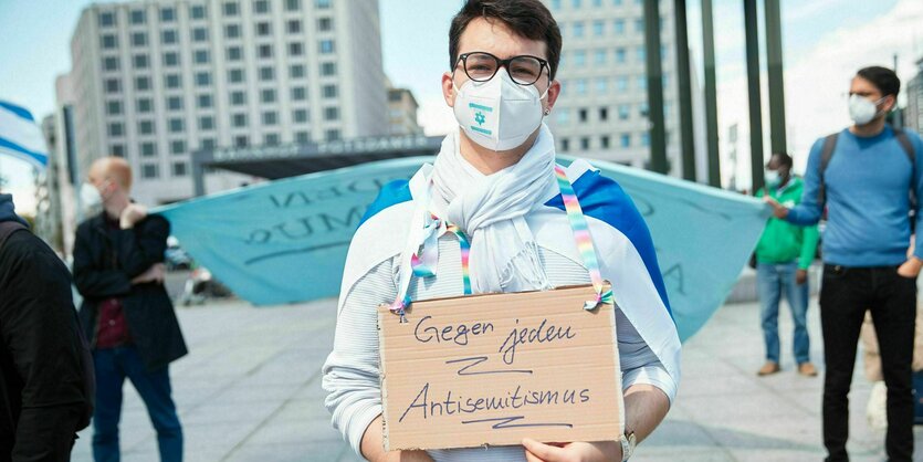 Ein Mensch trägt eine Maske und hält ein Schild hoch, auf dem steht: "Gegen jeden Antisemitismus"