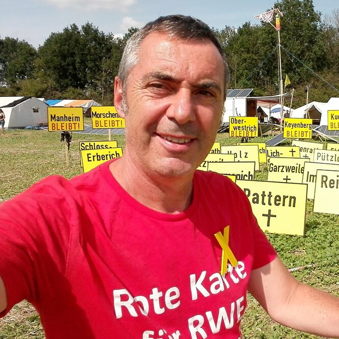 Porträt von Markus Dufner im roteh Shirt mit Aufschrift "Rote Karte für RWE" vor gelben Protestplakaten auf grüner Wiese