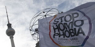 Links im Bild ist der Fernsehturm zu sehen, rechts eine weiße Flagge, auf der steht "Stop Homophobia" und in kleineren Buchstaben "https://enough-is-enough.eu". Im Hintergrund der Flagge sieht man einen Teil der Weltzeituhr