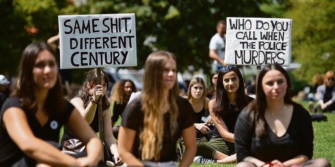 Demonstrantinnen sitzen im Gras und halten Schilder hoch.