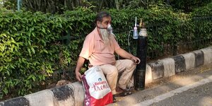 A man sitting on a kerbstone. He is middle aged with a long beard and wears an oxygen mask.