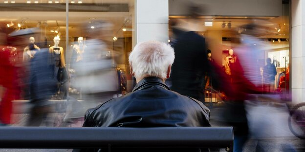 ein Rentner sitztauf einer Bank in einer Einkaufsstrasse