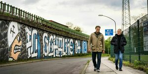 Olivier Kruschinski und Bodo Menze gehen an einer Mauer mit großem Graffiti vorbei: FC Schalke 04