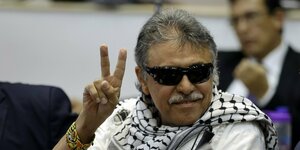 Jesús Santrich, ein grauhaariger Mann mit dunkler Brille und Palästinensertuch, lächelt in die Kamera und macht das Victory-Zeichen