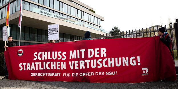 Menschen halten ein großes rotes Banner vor einem Gebäude. Aufschrift: "Schluss mit der staatlichen Vertuschung"