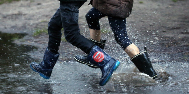 Zwei Kinder laufen in Gummistifeln durch Wasser auf der Straße