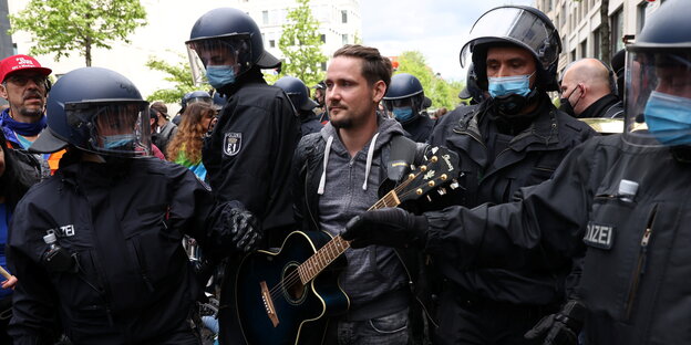 Ein junger Mann mit Gitarre und Langen Haaren, der von mehreren Polizisten abgeführt wird.