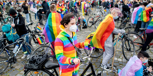 In Regenbogenfahnen gehüllte Radfahrer warten im Demozug darauf, dass es weitergeht.