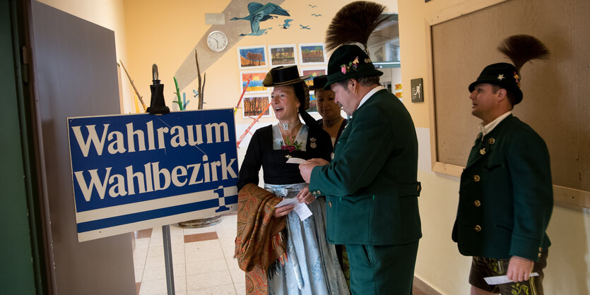 Menschen in Tracht-Kleidung stehen in einem Raum, auf einem Schild steht groß geschrieben: "Wahlraum"