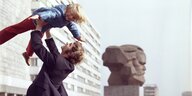 Erwachsene heben Kind hoch, vor dem Nischel-Denkmal in Chemnitz, ehemals Karl-Marx-Stadt