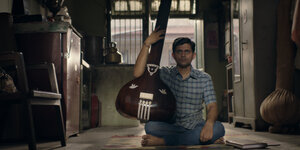Ein junger Mann sitzt mit seinem Instrument auf dem Boden zwischen den Möbeln
