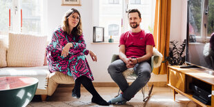 Ein Mann und eine Frau sitzen in einem Wohnzimmer. Im Hintergrund die Fenster mit Tageslicht