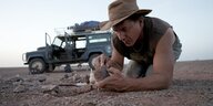 Mann am Boden untersucht Gestein - hinter ihm ein Jeep