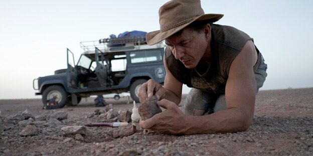 Mann am Boden untersucht Gestein - hinter ihm ein Jeep