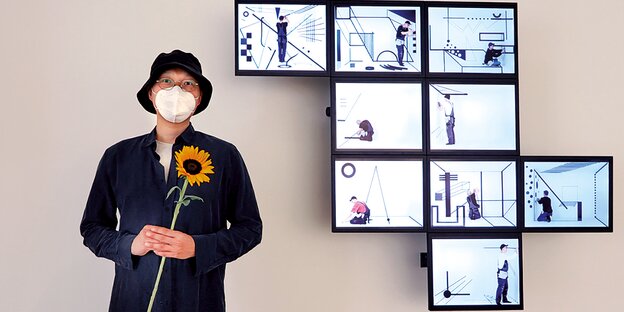 Der Künstler Hyunho Park steht vor seiner Videoinstallation aus neun Bildschirmen. Er hält eine Sonnenblume in der Hand.