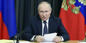 Wladimir Putin sitzt zwischen russischen Flaggen vor einem Mikrofon