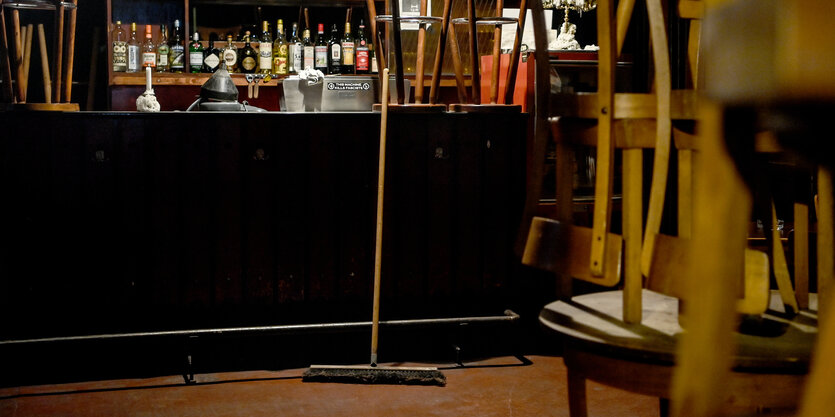 Blick durch ein Fenster in eine geschlossene Kneipe im Schillerkiez, in der ein Besen an der Bar lehnt. Hinter dem Tresen steht ein Regal, indem viele volle Alkoholflaschen stehen.