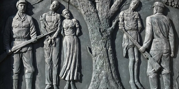 Das Denkmal zeigt Zwei Soldaten in Uniform und mit Gewehren. Sie stehen vor einem Baum an dem drei Menschen aufgehängt sind.