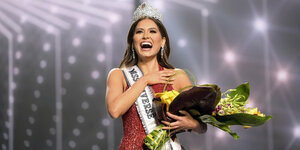 Eine Frau in einem roten Kleid, mit einer Tiara, einem großen Blumenstrauß und einer Schärpe, auf der steht: Miss Universe
