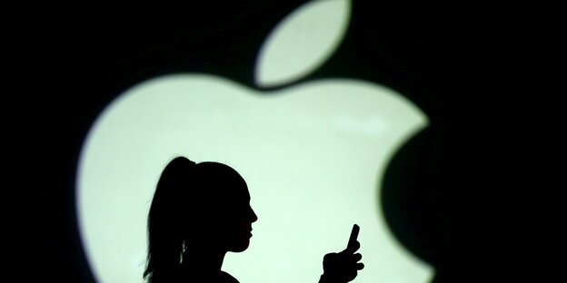Eine Person hält ein Handy in der Hand. Im Hintergrund ist das Logo der Firma Apple zu sehen
