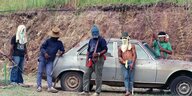 Fünf Männer mit Mützen oder Tüchern vor dem Gesicht und Gewehren stehen um einen Wagen