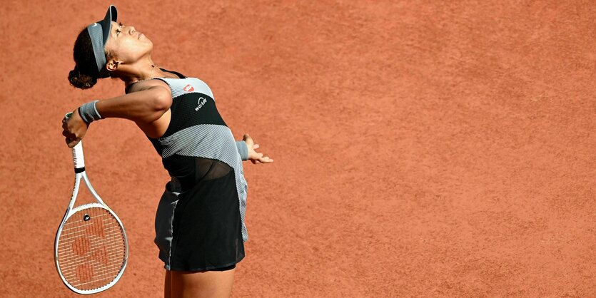 Eine Frau steht allein auf einem Platz und blickt nach oben. In ihrer rechten Hand hält sie einen Tennisschläger, den sie über die Schulter zu einem weiten Schlag ausgeholt hat.