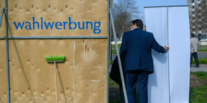 Im Vordergrund die Rückseite eines Wahlplakats. Kandidat Tobias Krull, mit dem Rücken zur Kamera, stützt ein weiteres Plakat