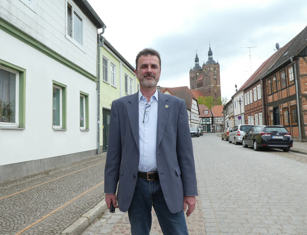 Rüdiger Kloth steht auf einer Straße. Im Hintergrund sind niedrige Wohnhäuser und eine Kirche. Kloth trägt einen Anzug und hat seine Brille ins Hemd gesteckt. Er ist ein mann mittleren Alters mit kurzen Haaren und Dreitagebart. Rüdiger Kloth steht auf einer Straße. Im Hintergrund sind niedrige Wohnhäuser und eine Kirche. Kloth trägt einen Anzug und hat seine Brille ins Hemd gesteckt. Er ist ein mann mittleren Alters mit kurzen Haaren und Dreitagebart.