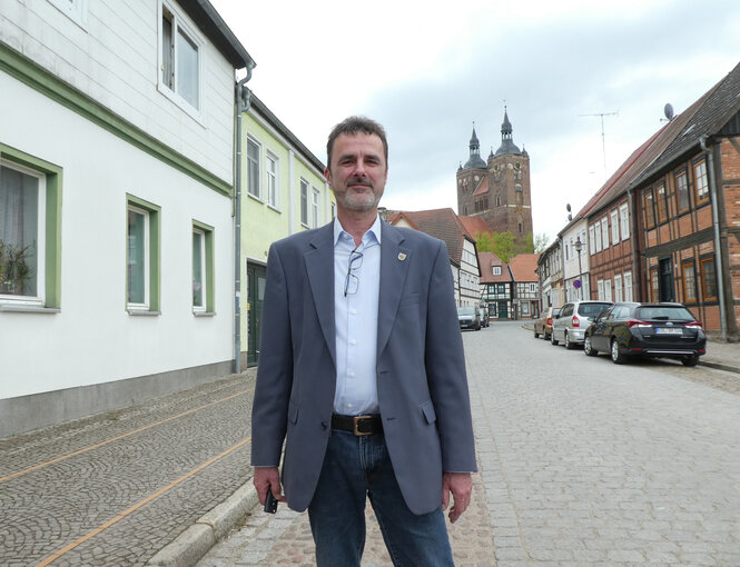 Rüdiger Kloth steht auf einer Straße. Im Hintergrund sind niedrige Wohnhäuser und eine Kirche. Kloth trägt einen Anzug und hat seine Brille ins Hemd gesteckt. Er ist ein mann mittleren Alters mit kurzen Haaren und Dreitagebart.