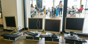 Mehrere Computer und Monitore auf einem Tisch in einer Schule - im Hintergrund, SchülerInnen und Lehrer