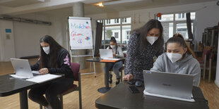 Junge Frauen sitzen an Einzeltischen hinter Laptops, eine ältere Frau betreut sie.