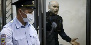 Andrej Piwowarow gestikuliert hinter einer Glasscheibe vor Gericht - auf der anderen Seite ein Polizeibeamter mit Gesichtsmaske