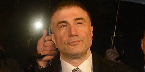 Mafia-Boss Sedat Peker, ein Mann im Anzug-hinter ihm Scheinwerferlicht, neben ihm Männer, die Regenschirme über ihn halten