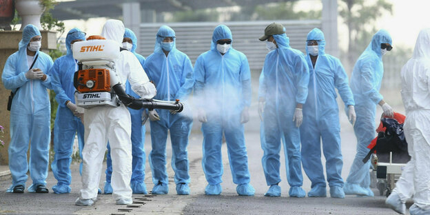 Eine Gruppe von Personen in blauen Schutzanzügen wird von einer Person in einem weißen Schutzanzug mit Desinfektionsmittel besprüht Eine Gruppe von Personen in blauen Schutzanzügen wird von einer Person in einem weißen Schutzanzug mit Desinfektionsmittel besprüht