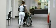 Eine junge Frau schiebt einen älteren Mann im Rollstuhl durch ein Besuchszimmer